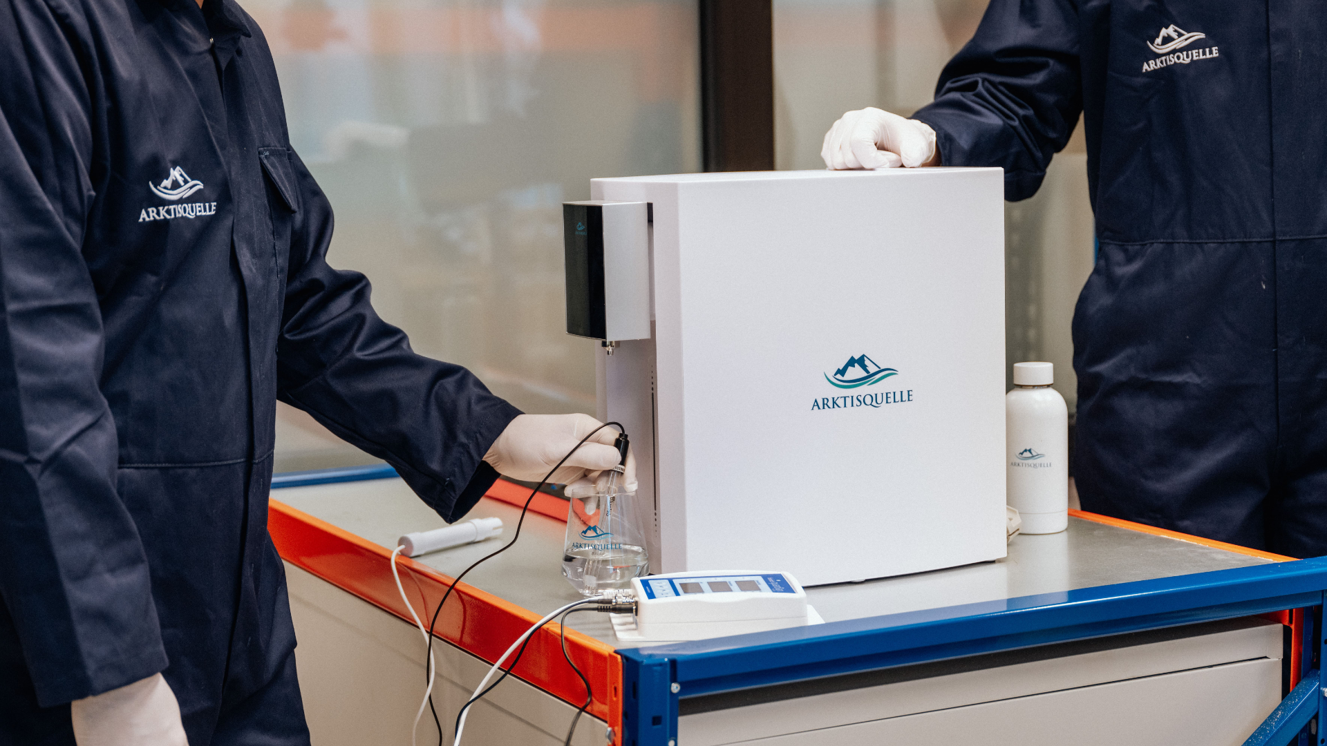 Zwei Techniker in Arktisquelle-Montagekleidung prüfen die Wasserqualität einer weißen Arktisquelle Osmoseanlage mithilfe eines elektronischen Messgeräts – Qualitätskontrolle im Fertigungsbereich.