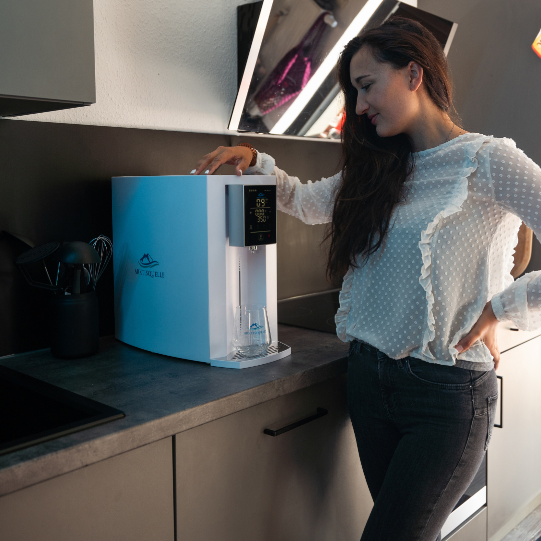 Junge Frau in weißer Bluse bedient lächelnd eine weiße Arktisquelle Osmoseanlage mit digitalem Display in einer modernen Küche – stilvolle Wasserfilterstation mit Trinkglas.