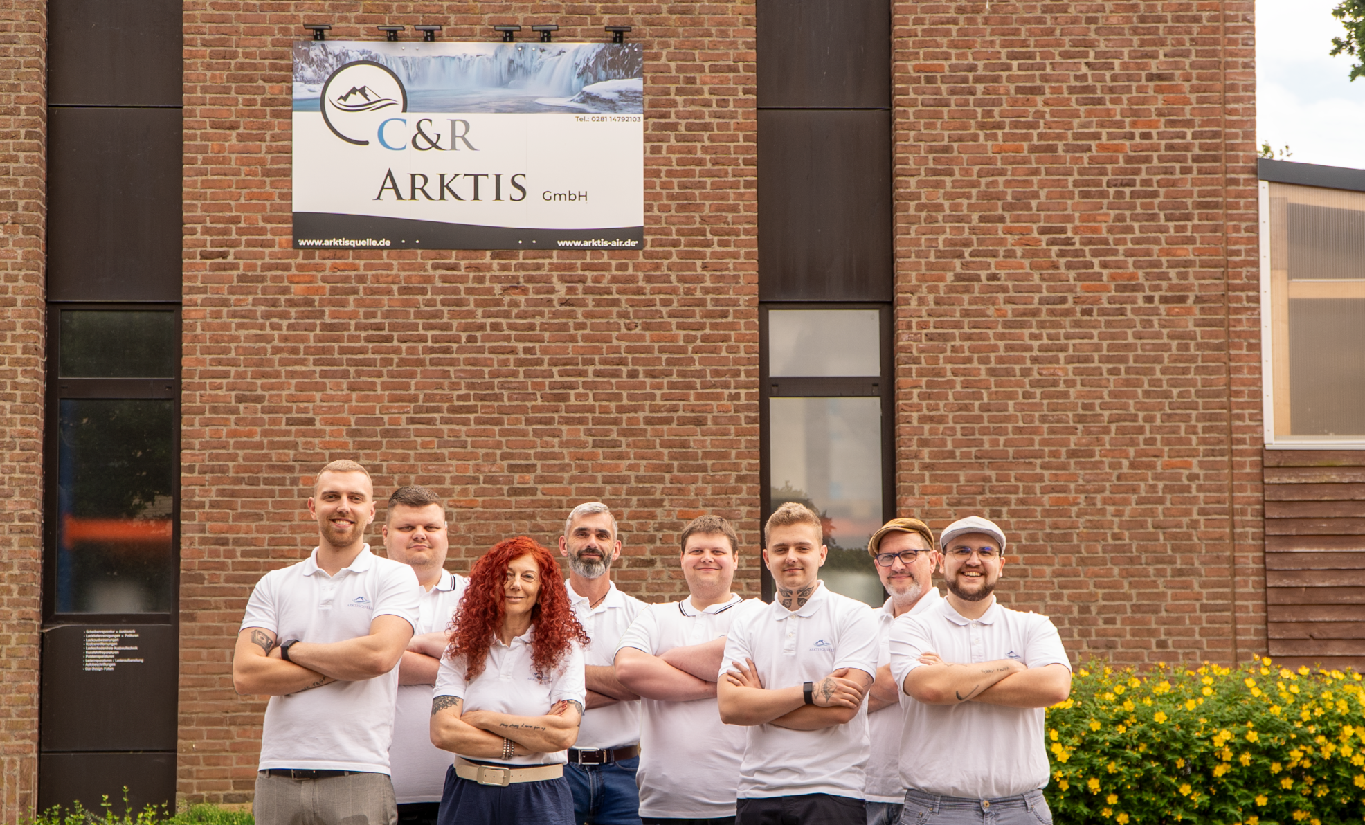 Teamfoto der Arktisquelle vor dem Firmengebäude der C&R Arktis GmbH mit Backsteinfassade – zehn Mitarbeitende in weißen Polo-Shirts posieren lächelnd mit verschränkten Armen unter dem Firmenschild.